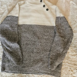 Stickad tröja från DEVERED 1902 - Säljer en snygg stickad tröja från DEVERED 1902 i storlek L. Tröjan är tvåfärgad med vit upptill och gråmelerad nedtill, har lång ärm och hög krage med tre dekorativa knappar på axeln. Perfekt för kyliga dagar.