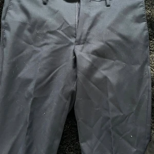 Blåa kostymbyxor från J Lindberg - Snygga blåa kostymbyxor med klassisk rak passform och hällor för bälte. Byxorna har diskret dragkedja framtill och är tillverkade i ett slätt tyg som ger en stilren look. Perfekta för dig som gillar en clean och enkel stil.