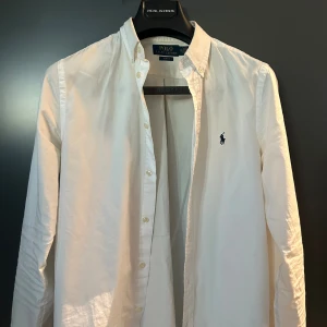 Vit skjorta från Polo Ralph Lauren Strl 170  - Klassisk vit skjorta från Polo Ralph Lauren i slim fit-modell. Skjortan har långa ärmar, knappar framtill och den ikoniska broderade loggan på bröstet. Tillverkad i mjuk bomull och passar perfekt till en stilren look. Ganska redigt använd men fortfarande fint skick! 