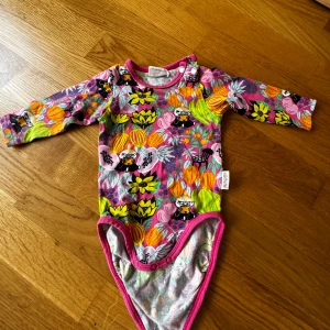 Färgglad Mumin-body från Martinex - Superfin långärmad body från Mumin by Martinex i storlek 62. Bodyn har ett färgstarkt blommigt mönster i rosa, gul, orange och grön med små Mumin-karaktärer. Tillverkad i mjuk bomull och har tryckknappar vid axeln och i grenen för enkel påklädning.
