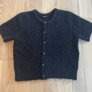 Mörkblå kofta från Gina Tricot - Mysig mörkblå kofta från Gina Tricot med kort ärm och fluffig, stickad känsla. Koftan har rund halsringning och snygga guldfärgade knappar framtill.