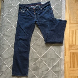 Blå raka jeans från Diesel, stl 32 - Säljer ett par klassiska blå jeans från Diesel i rak modell. Jeansen har fem fickor, knappgylf och snygga detaljer på bakfickorna. Materialet är 93% bomull, 5% polyester och 2% elastan för en skön passform.
