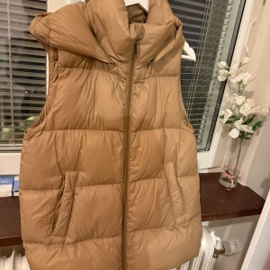 Beige dunväst med hög krage - Snygg beige dunväst med quiltad design och hög krage. Västen har två stora fickor framtill och stängs med dragkedja. Perfekt för lager-på-lager och ger en cool, avslappnad look.