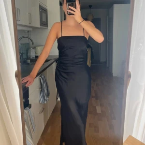 Svart lång satinklänning med smala band - Säljer en svart långklänning i satin med smala axelband och figurnära passform. Klänningen har en stilren, minimalistisk look och lätt rynkade detaljer längs sidan för en snygg siluett. Perfekt för dig som gillar en elegant och enkel stil.ALLDRIG ANVÄNTS