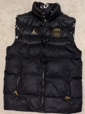 Svart dunväst PSG x Jordan, vändbar - Vändbar svart dunväst från Jordan x PSG med coolt Paris-tryck på ena sidan och diskret PSG-logga samt Jumpman på andra. Västen har dragkedja, två fickor med gulddetaljer och hög krage. Perfekt för dig som vill sticka ut med sportig streetstyle. Vändbar ut o in. pris kan diskuteras
