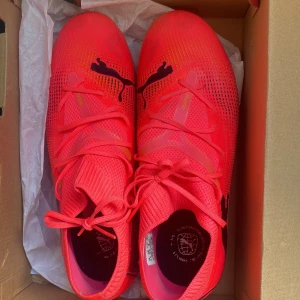 Puma neonrosa fotbollsskor med dobbar - Säljer ett par grymma neonrosa Puma fotbollsskor med svarta detaljer och coolt mönster på sidan. Skorna har snörning, plös i stickad stil och dobbar i rosa och gult för bästa grepp på planen. Perfekta för dig som vill sticka ut på matchen!