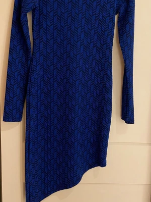 Blå långärmad mönstrad klänning - Snygg blå klänning med svart grafiskt mönster och lång ärm. Klänningen har en asymmetrisk nederkant och är tillverkad i ett mjukt, stretchigt material som sitter bekvämt. Perfekt för dig som gillar unika detaljer och vill sticka ut.