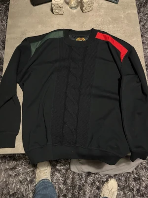 Svart tröja USA Marine med kabelstickning - Svart långärmad tröja från USA Marine med bred kabelstickad panel framtill. Har gröna och röda detaljer på axlarna och rund hals. Märkespatch med broderad logga på insidan. Perfekt för dig som gillar streetwear med lite extra edge.