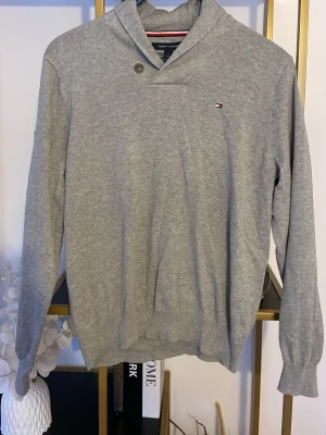 Grå tröja från Tommy Hilfiger - Säljer en stilren grå tröja från Tommy Hilfiger med liten logga på bröstet. Tröjan har en unik krage med knappdetalj och är tillverkad i mjuk bomull. Långärmad modell med ribbade muddar vid ärmslut och nederkant. Perfekt till höst och vår.