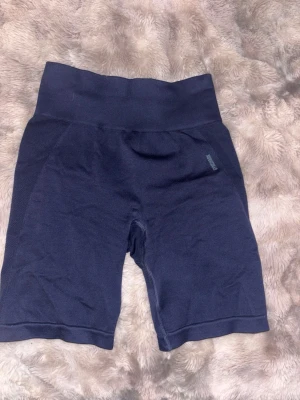 Gymshark shorts - Säljer ett par mörkblå biker shorts från Gymshark med hög midja och bred resår där loggan syns tydligt baktill. Materialet är stretchigt och syntetiskt, perfekt för träning. Snygg, tight passform som sitter skönt och följer kroppen.