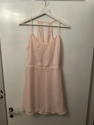 Ljusrosa kort klänning från Bershka - Supersöt ljusrosa klänning från Bershka med tunna axelband och snygg öppen ryggdetalj. Klänningen är kort och har en lätt, flowy passform i tunt tyg som känns luftigt. Perfekt för varma dagar och somriga vibes!