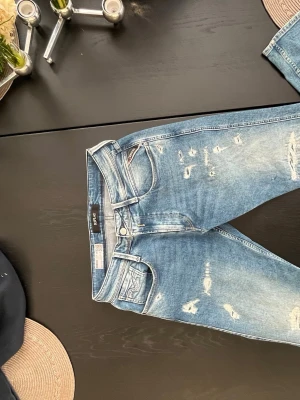 Replay blå slitna jeans - Säljer ett par blå jeans från Replay Anbass med snygga slitningar och detaljer på både fram- och baksida. Klassisk femficksmodell med ljus tvätt och bruna läderpatch bak i midjan. Perfekt för dig som gillar en cool och avslappnad stil. 