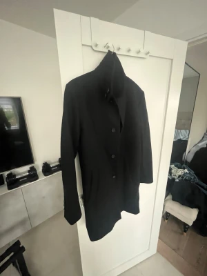 Svart mörkblå med knappar, Tiger of Sweden  - Stilren mörkblå  rock med klassisk krage och tre knappar framtill. rocken har en rak och lite längre passform som ger en snygg siluett. Perfekt för dig som gillar en clean och enkel look. Materialet känns mjukt och passar bra till kyligare dagar. Storleken på rocken är S