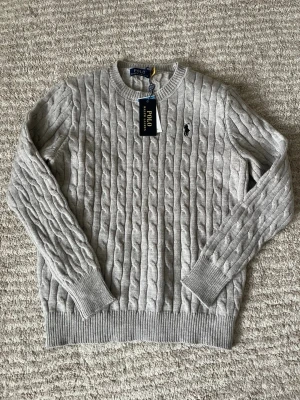 Grå kabelstickad tröja Polo Ralph Lauren - Snygg grå kabelstickad tröja från Polo Ralph Lauren med rund hals och klassisk broderad logga på bröstet. Tröjan har långa ärmar och ribbade muddar vid ärmslut och nederkant. Perfekt för dig som gillar stilrena och tidlösa plagg.