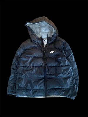 Svart Nike pufferjacka med huva - Säljer en svart pufferjacka från Nike med Therma-Fit och stor huva. Jackan har dragkedja framtill, två sidofickor och ett stort vitt Nike-logga på bröstet. Materialet är glansigt och känns lätt men ändå varmt, perfekt för kyliga dagar. Kolla bilder för skick