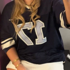 Blå fotbollströja med nummer 17 - Snygg marinblå fotbollströja med stora vita siffror 17 på framsidan. Tröjan har korta ärmar med vita ränder och mesh-detaljer nedtill för extra ventilation. V-ringad hals med vit kant. Tröjan är från urban outfitters endast använd en gång 