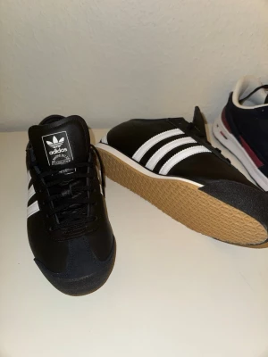 Adidas Italia svarta sneakers - Säljer ett par Adidas Italia sneakers i svart med klassiska vita ränder och gummisula. Skorna har snörning, rund tå och är tillverkade i skinn och syntet. Snygg retrostil med Adidas-logga på plösen och sidan. Perfekt för dig som gillar ikoniska modeller.