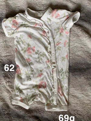 Vit blommig kortärmad pyjamas - Söt vit pyjamas från Newbie med korta ärmar och blommigt mönster i rosa och grönt. Pyjamasen har knappar framtill och en liten volang längs knappraden. Perfekt för varma dagar och mjuk mot huden.🌸 Fint använt skick. Storlek 62