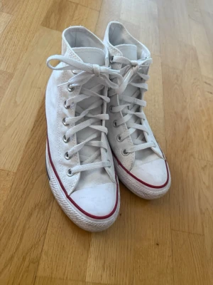 Vita Converse All Star high-top sneakers - Klassiska vita Converse All Star high-top sneakers med röd rand längs sulan och rund tå. Skorna har snörning och är tillverkade i canvas med gummisula. Perfekta för dig som gillar en tidlös och clean stil.