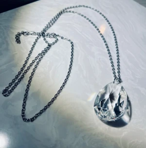 Halsband med stor droppformad kristall - Unikt halsband med en lång silverfärgad kedja och en stor, droppformad kristall som hänge. Kristallen är slipad för att reflektera ljuset och ger ett riktigt glittrigt intryck. Perfekt för dig som vill sticka ut med ett statement-smycke.