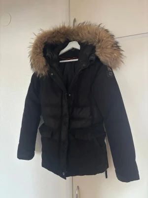 Svart parkajacka med päls från Hollies - Parkajacka från Hollies i svart med fluffig päls runt huvan. Jackan har långa ärmar, stora fickor och snygga detaljer som knappar och dragkedja. Perfekt för kalla dagar och har en klassisk look med badge på ärmen.