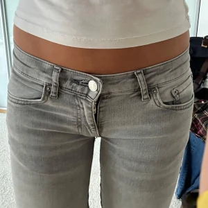 Grå low waist bootcut jeans  - Säljer ett par ljusgrå jeans med bootcut passform och låg midja. Jeansen har klassisk femficksdesign, dragkedja och knapp framtill. Omsydda och lite slitningar vid benkanten och där benen delar på sig pga omsyningen.