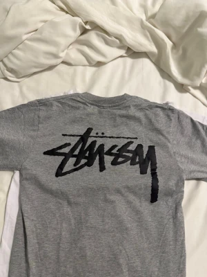 Grå Stüssy t-shirt med logga - Säljer en grå t-shirt från Stüssy med stor svart logga på ryggen och mindre logga på bröstet. Klassisk rund hals och korta ärmar. Materialet är mjuk bomull som är skönt mot huden. Perfekt för dig som gillar streetwear och vill ha en clean look.