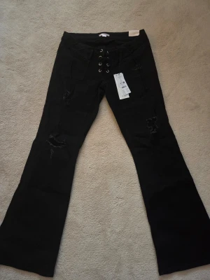 Svarta bootcut jeans från Gina Tricot - Bootcut jeans i svart från Gina Tricot, modell 14+ pleated tie jeans. Låg midja och snörning framtill med metallöljetter. Råa slitningar på benen ger en edgy vibe. Materialet är klassisk jeans och passformen är loose. Perfekt för dig som gillar statement-plagg.