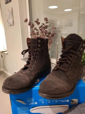 Bruna mockaboots från Replay 42 - Snygga bruna boots i mocka från Replay med snörning framtill och dragkedja på sidan. Skorna har rund tå och platt sula, samt detaljerade sömmar och en robust känsla. Perfekta för dig som gillar en stilren och avslappnad look.