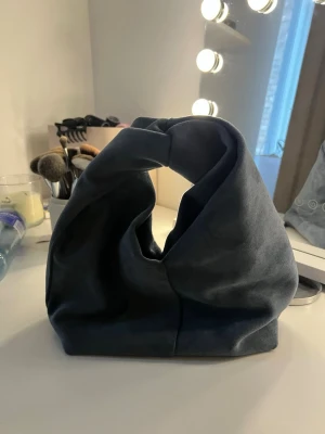 mocka handväska - Säljer en stilren handväska i mjuk mocka med unik knytning upptill. Väskan har ett magnetlås och är rymlig inuti, perfekt för att bära med sig det viktigaste. Enkel design utan mönster eller logga, passar dig som gillar minimalistiskt.