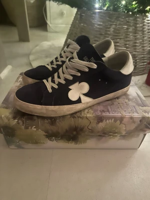 Mörkblå sneakers från The Gallerist - Säljer ett par mörkblå sneakers från The Gallerist i mocka med vit snörning och vit klöverdetalj på sidan. Skorna har vit häl med logotyp och mönstrad rem med klöver på sidan.   