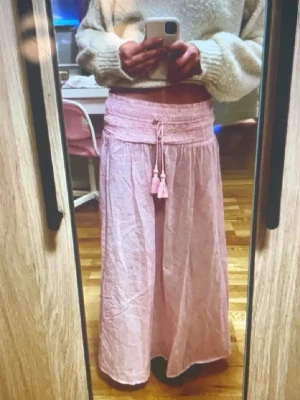 Ljusrosa långkjol med snörning - Säljer en ljusrosa långkjol med smockad midja och snörning med pärldetaljer och tofsar. Kjolen har ett svagt paisleymönster och är gjord i ett tunt, flowy material som känns luftigt. Perfekt för sommaren. 