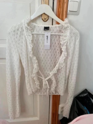 Vit omlott blus med volang från Gina Tricot - Säljer en vit omlott blus från Gina Tricot i storlek M. Blusen har lång ärm, volangdetaljer längs halsringningen och framtill, samt knyt framtill. Materialet är lätt och har ett diskret geometriskt mönster. Perfekt för dig som gillar romantiska detaljer. Aldrig använd och säljer 100 kronor billigare!