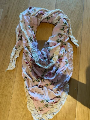 Blommig rosa sjal med spetskant - Säljer en romantisk trekants sjal i ljusrosa med blommigt mönster och dekorativ spetskant. Materialet känns lätt och luftigt, och blommorna går i grönt, vitt och rosa. Från Ester Elenora 