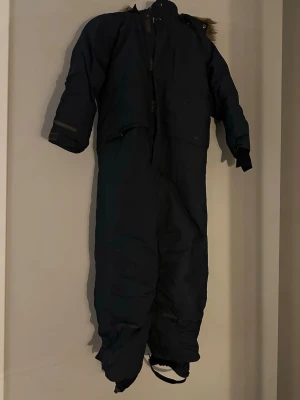 Mörkblå vinteroverall Didriksons 130 - Säljer ett par mörkblå täckbyxor från Didriksons i storlek 130. Byxorna är 100% vattentäta och har avtagbar huva med fuskpäls, reflexdetaljer och justerbara benslut med fotstrappar. Perfekta för kalla vinterdagar och snölek.