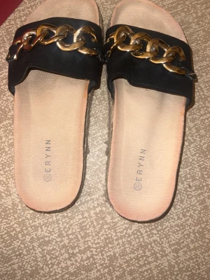 Svarta sandaler med guldkedja från Erynn - Säljer ett par svarta sandaler från Erynn med bred rem i mocka och en stor guldfärgad kedjedetalj. Sulan är platt och har en beige flätad kant som ger en chill vibe. Perfekta för sommardagar och enkla att glida i. Aldrig använda