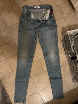 Tommy Jeans ljusblå jeans - Säljer ett par ljusblå jeans från Tommy Jeans med klassisk femficksdesign och diskret logga på bakfickan. Jeansen har dragkedja och knapp framtill, samt bälteshällor. Materialet är mjukt denim i bomull med lite stretch för skön passform.