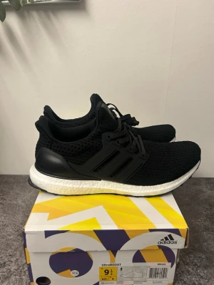 Adidas UltraBoost svarta sneakers - Svarta Adidas UltraBoost sneakers med stickad ovandel och klassiska tre ränder på sidan. Skorna har snörning, vit Boost-mellansula och svart yttersula med Continental-gummi. Perfekta för dig som gillar stilren och sportig look.