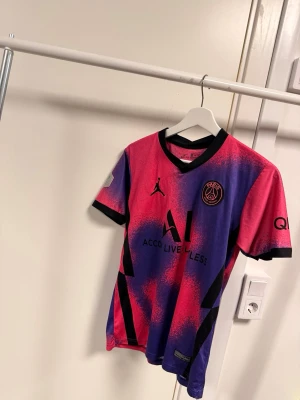 PSG Mbappé matchtröja rosa/lila - Säljer en PSG matchtröja med Mbappé #7 på ryggen. Tröjan har en fet rosa och lila färg med svart krage och detaljer, samt PSG-logga och Jordan-logga på bröstet. Materialet är lätt polyester som andas, perfekt för fotboll eller träning.