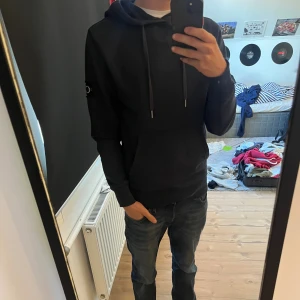Blå hoodie från Calvin Klein - Mörklblå hoodie från Calvin Klein med klassisk huva och snörning. Stor CK-logga på vänster ärm ger en clean och modern vibe. Mjuk bomullskänsla och känguruficka framtill. Perfekt för chill dagar eller när du vill ha en stilren streetlook.
