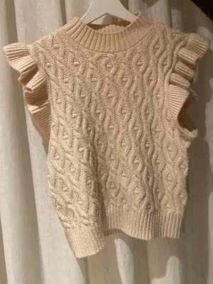 Stickad tröja med volang - Supersöt beige stickad väst från Name It med flätmönster och volangdetaljer vid axlarna. Säljer då den tyvärr är för liten. Använd fåtal gånger.