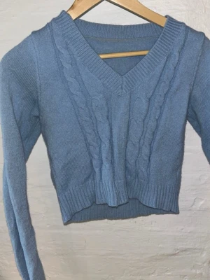 Blå croppad stickad tröja - Säljer en blå croppad stickad tröja med flätstickade detaljer framtill och v-ringad hals. Tröjan har långa ärmar och bred ribbad mudd vid hals, ärmslut och nederkant. Perfekt för dig som vill ha en mysig men trendig look.