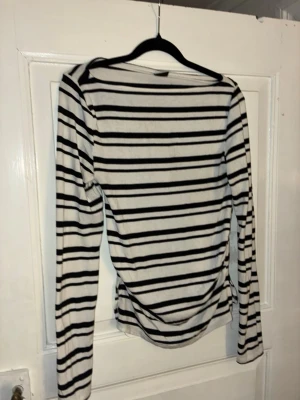 Randig långärmad topp i bomull - Säljer en vit långärmad topp med svarta horisontella ränder. Toppen har en enkel, rak passform och är gjord i mjuk bomull. Perfekt till jeans eller kjol för en klassisk look.