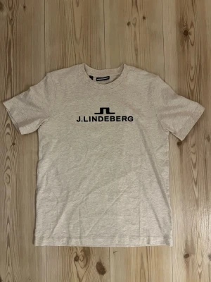 Beige t-shirt från J.Lindeberg XS - Snygg beige t-shirt från J.Lindeberg i mjuk bomull. Klassisk passform med rund hals och svart logotyptryck framtill. Perfekt till jeans eller shorts för en clean och enkel stil.