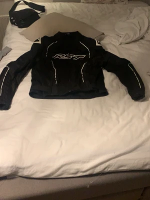 Svart mc RST jacka  - Säljer en svart skinnjacka från RST med vita detaljer och logga på bröstet. Jackan har lång ärm och sportig passform, perfekt för dig som gillar en cool och edgy stil. Snygga vita linjer längs ärmar och axlar ger extra attityd. Rst mc jacka, mycket bra skick och allmänt en skön mc jacka för våren sommaren och hösten, storlek UK 42 eller M:1000kr Mc kläder Mc ställ