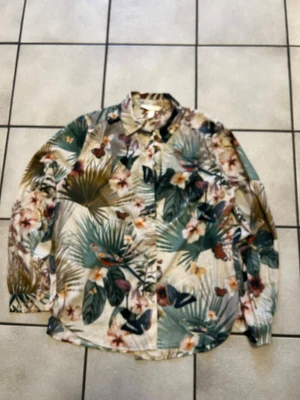 Blommig skjorta från H&M, strl 36 - Snygg skjorta från H&M i storlek 36 med tropiskt blommigt mönster och fjärilar i grönt, beige, brunt och inslag av orange. Skjortan har klassisk krage, långa ärmar och knappar framtill. Perfekt för dig som gillar färgglada och unika prints.