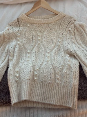 Beige stickad tröja med pärlor från Zara - Superfin beige stickad tröja från Zara i storlek S. Tröjan har ett snyggt kabelstickat mönster och är dekorerad med små pärlor över hela plagget. Rund hals och långa ärmar med ribbade muddar. Perfekt för dig som gillar detaljer och vill ha en mysig look.