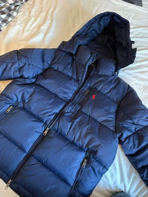 Blå pufferjacka från Polo Ralph Lauren - Säljer en blå pufferjacka från Polo Ralph Lauren i storlek M. Jackan har huva, två sidofickor med dragkedja och en röd broderad logga på bröstet. Perfekt för kalla dagar! Jackan är lite skrynklig du den precis varit nerpackad men det försvinner efter en kort stund. Pris kan diskuteras!