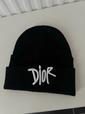 Svart Dior mössa med broderad logga - Säljer en svart mössa från Dior med vit broderad logga framtill. Mössan har en klassisk uppvikt kant och är tillverkad i mjukt stickat material. Perfekt för dig som vill ha en clean och stilren accessoar med lyxig känsla.