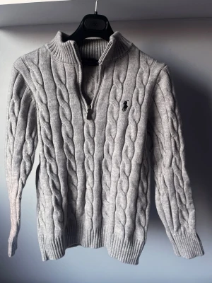 Grå kabelstickad tröja Polo Ralph Lauren - Kabelstickad grå tröja från Polo Ralph Lauren med halv dragkedja och hög krage. Klassisk logga broderad på bröstet. Tröjan är långärmad och har ribbade muddar vid ärmslut och nederkant. Perfekt för kyliga dagar.